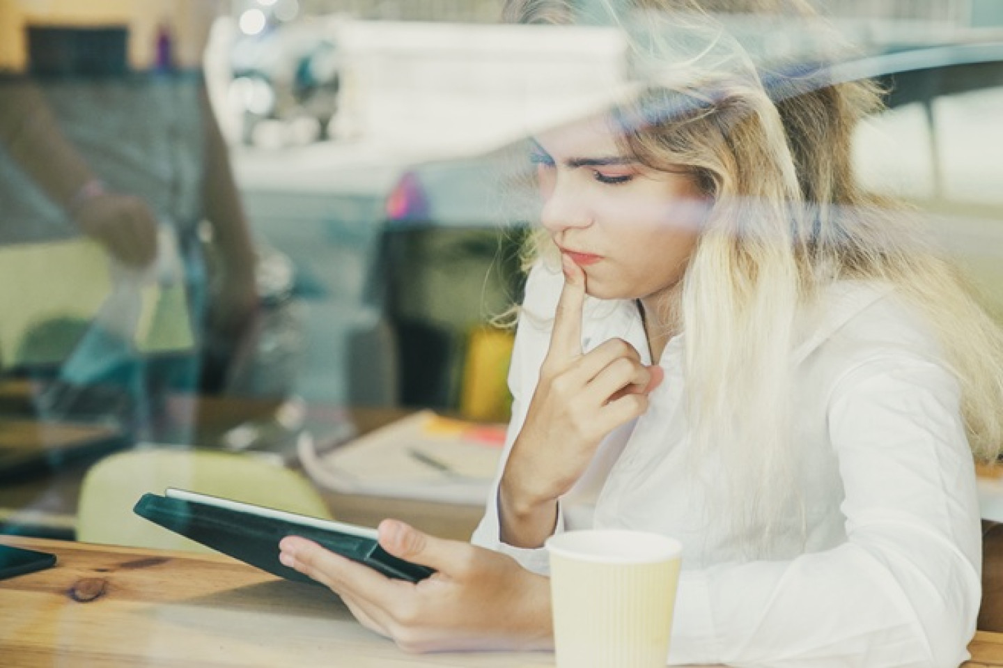 Pensive female professional sitting at desk in co-working space or coffee shop, using tablet. View from street through window glass. Communication concept pensamiento critico digital